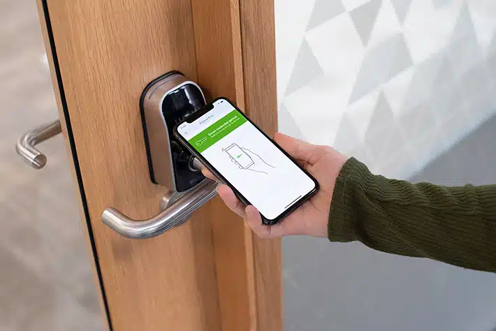 Mobiele telefoon met geopende toegangscontrole-app wordt tegen een digitaal deurslot gehouden, wat de eenvoudige en veilige sleutelloze toegang illustreert aangeboden door Meuleman’s toegangscontrolesystemen.